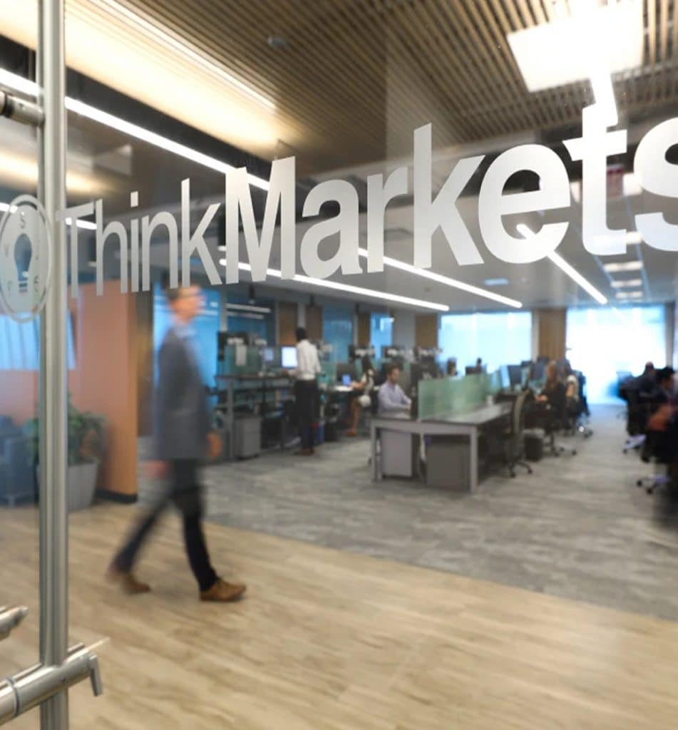 Interior view of a ThinkMarkets office showing an active trading floor with staff at work, viewed through a glass wall displaying the ThinkMarkets logo, representing a globally regulated online trading leader.